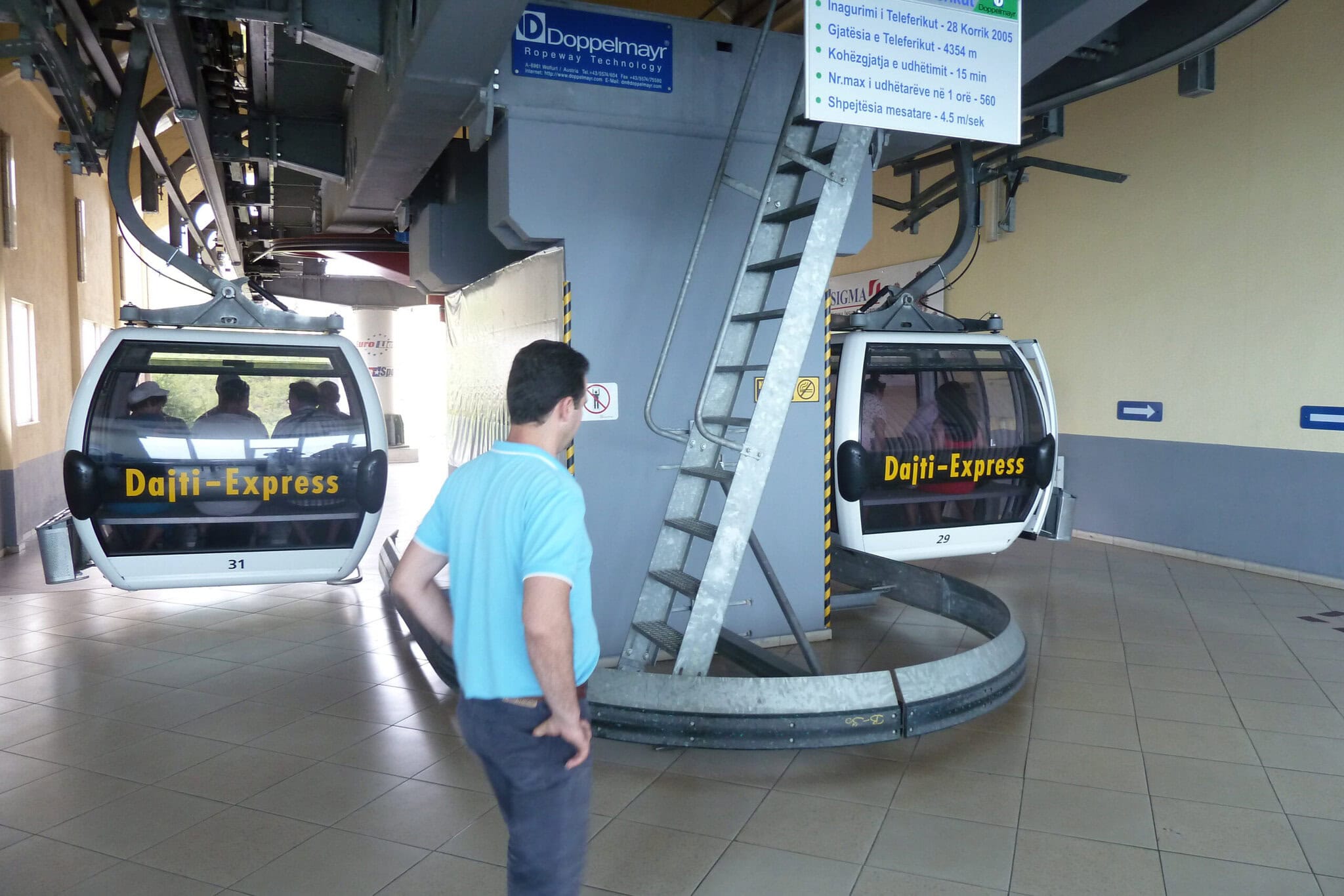 Two Dajti-Express gondola cabins at the lower station, with an informational sign showing the cable car's stats: opened 28 July 2005, 4,354 meters long, 15-minute ride