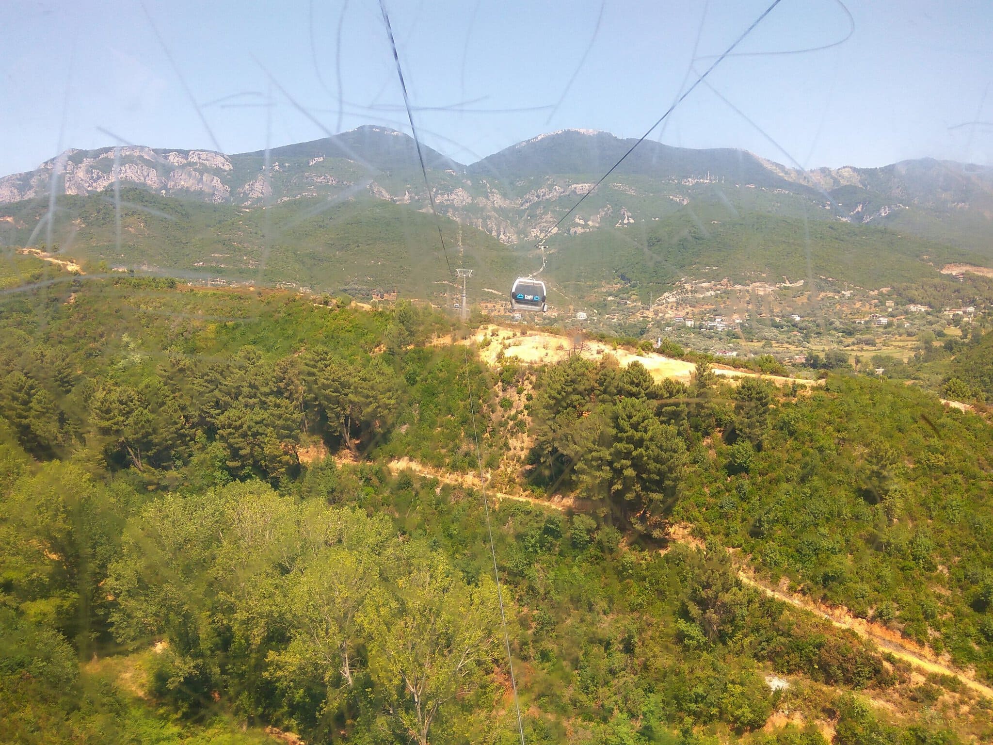 View from inside a Dajti Ekspres cabin mid-ride, looking out at another gondola crossing forested hills with limestone mountains in the background