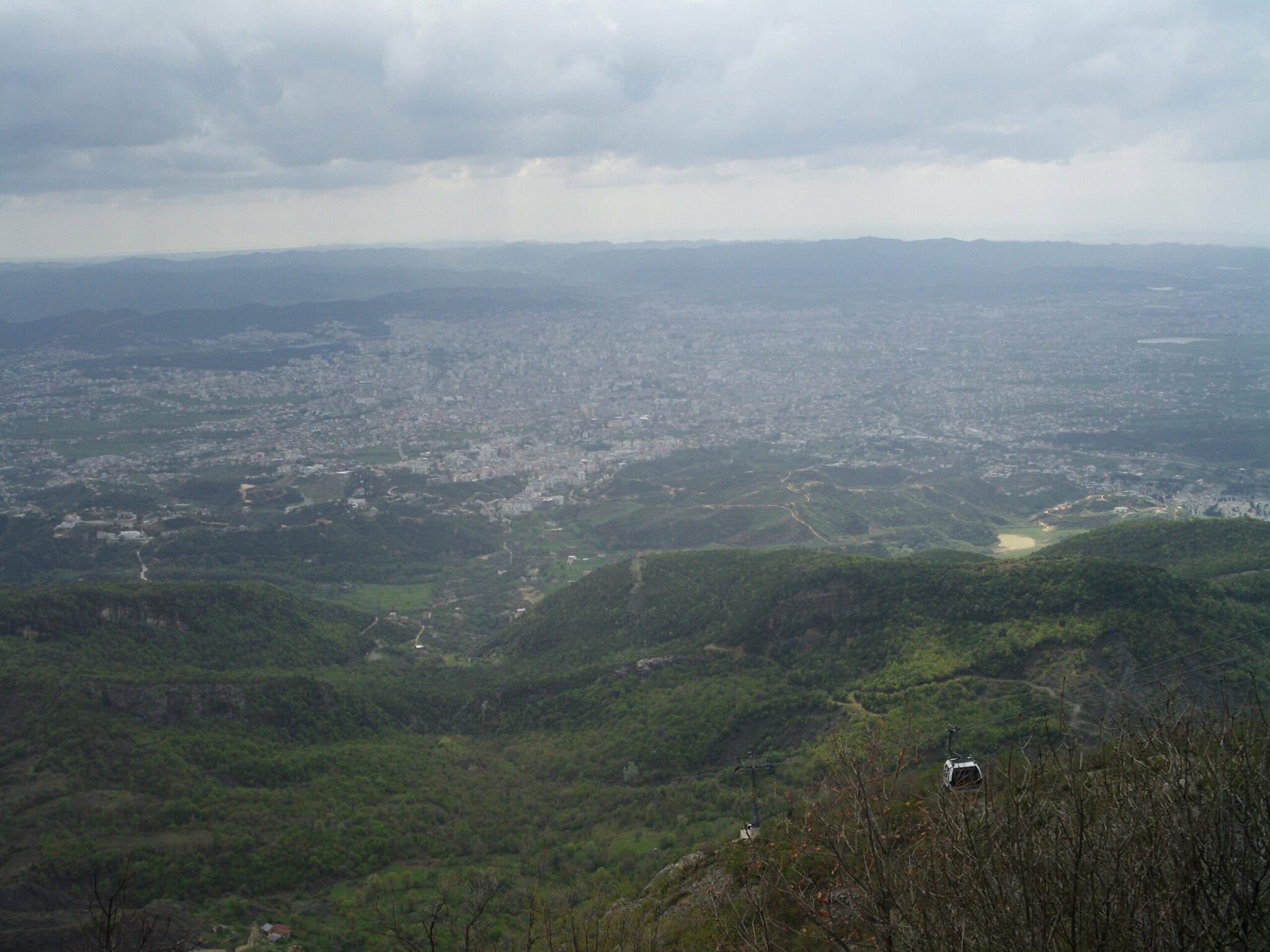 Panoramic view of Tirana from Mount Dajti, with a gondola cabin visible on the cable in the foreground and the city spreading out into hazy hills toward the Adriatic