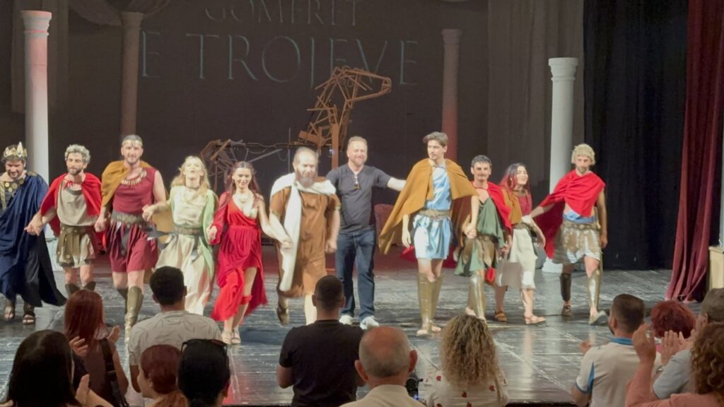 Full cast of Gomerët e Trojeve taking a curtain call at the Migjeni Theatre in Shkodra, with the director standing centre stage