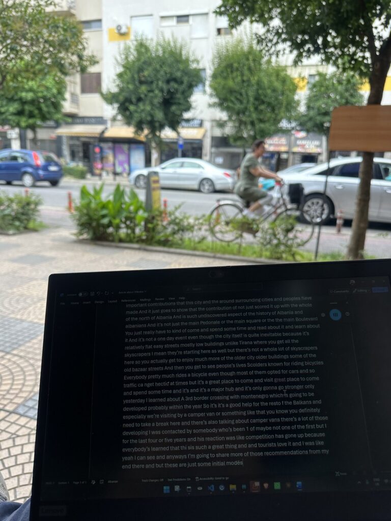 Laptop on a cafe table on a Shkodra street in the morning with a man cycling past in the background
